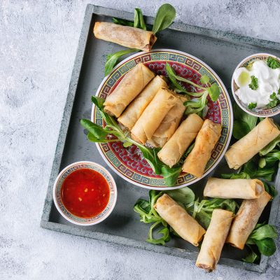 Fried spring rolls with red and white sauces, served in china plate on square wood tray with fresh green salad over gray blue texture background. Flat lay, space. Asian food
